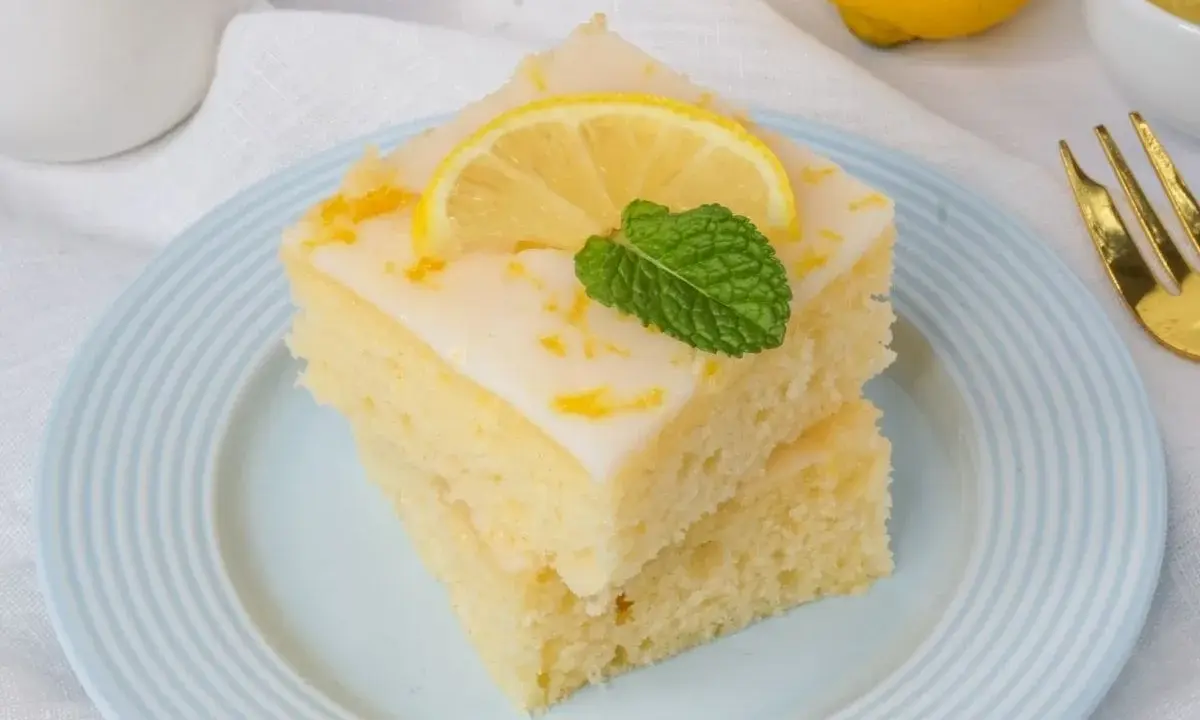 Zwei saftige Stücke weltbester Zitronenkuchen mit Joghurt, garniert mit Zitrone und Minze, auf einem blauen Teller.
