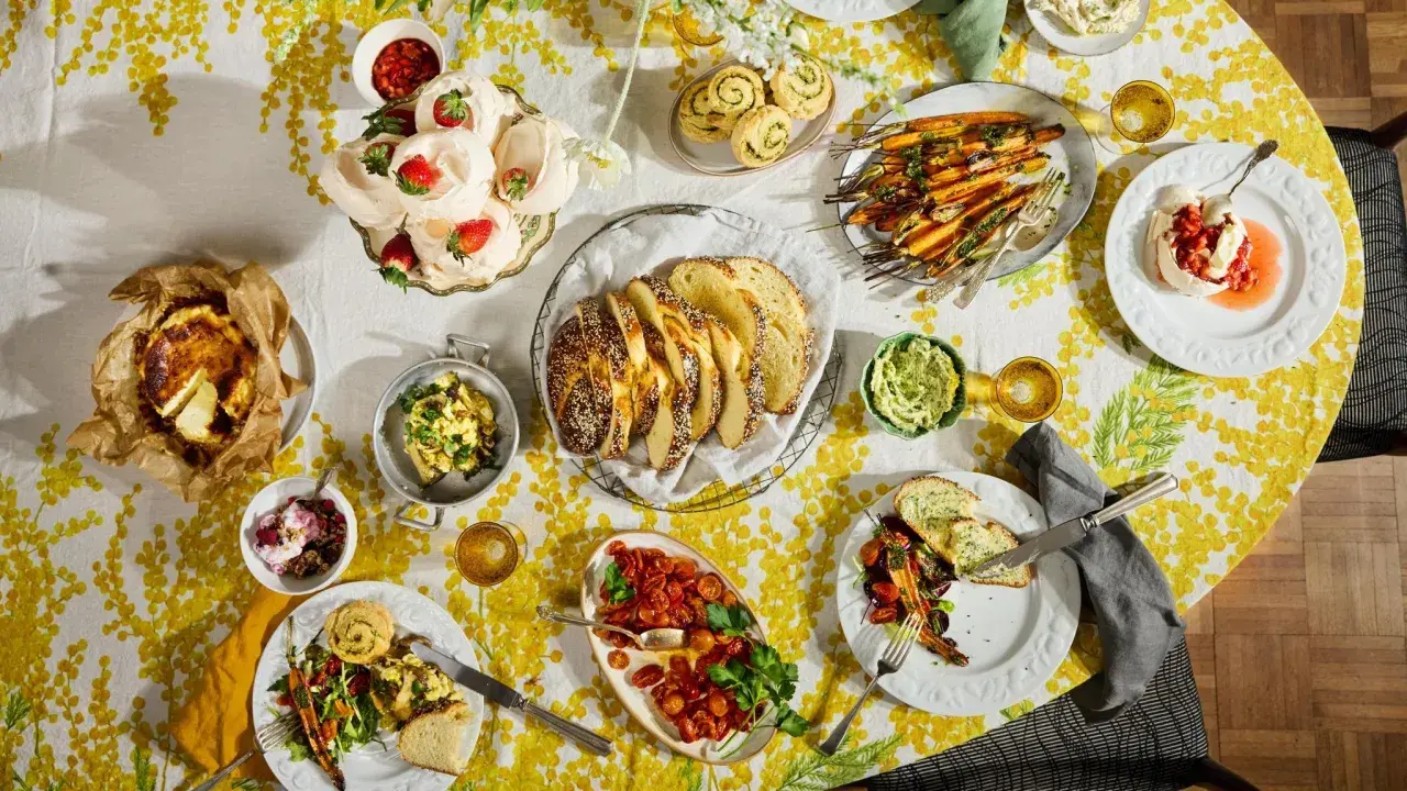 Tisch voller Oster-Essen-Ideen: Meringues mit Erdbeeren, Brot, Karotten, Quiche und mehr auf gelb-weißem Tischtuch.