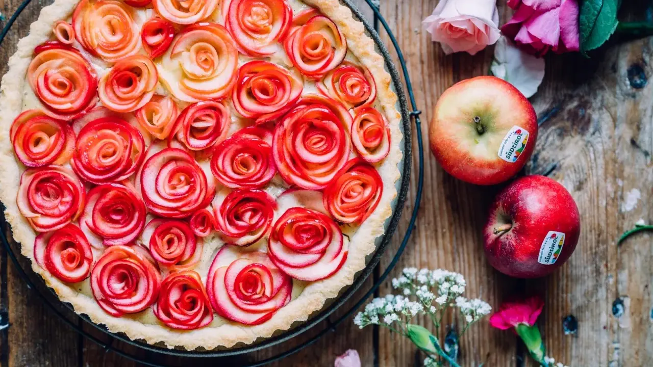 Ein wunderschöner Apfelrosen Kuchen, verziert mit zarten Apfelscheiben, die wie Rosen aussehen. Daneben liegen frische Äpfel und Blumen.