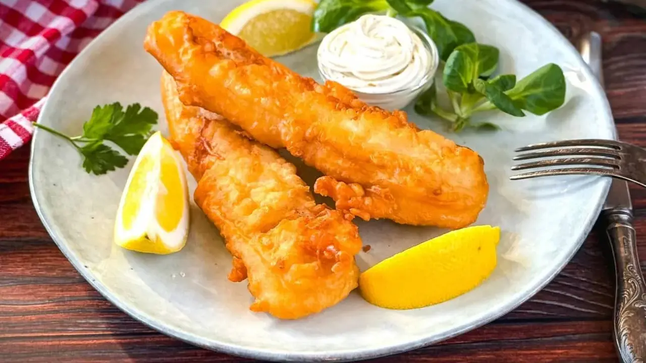 Zwei goldbraun frittierter Fisch mit Zitronenspalten, Sauce und Salat auf einem Teller.