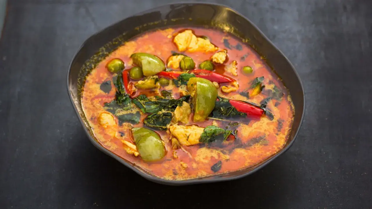 Ein dampfender rote thai curry mit Hühnchen, grünen Auberginen und roten Chilis in einer dunklen Schüssel.
