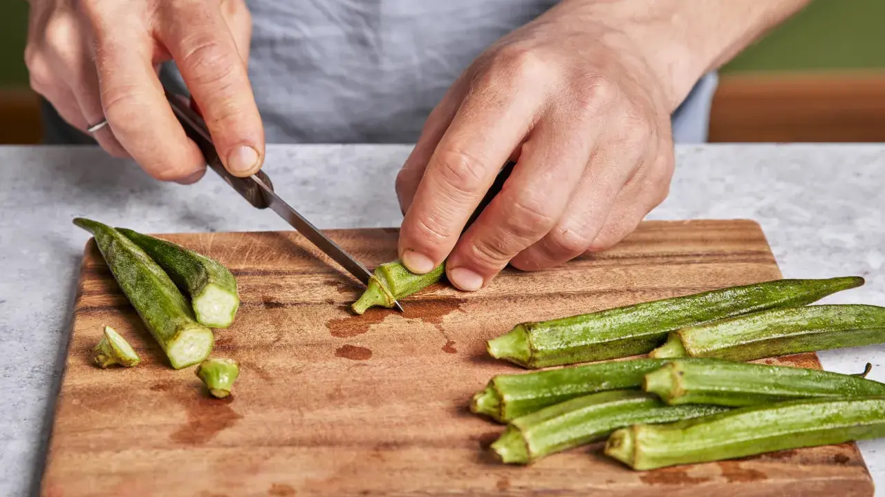 Hände schneiden Okra für ein leckeres Okra-Gericht. Frische grüne Schoten liegen auf einem Holzbrett.