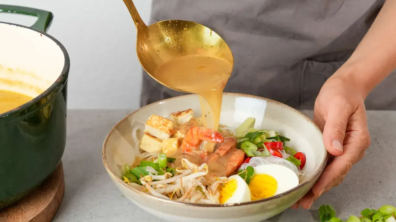 Eine goldene Schöpfkelle gießt eine cremige Brühe über Garnelen, Tofu, Ei und Sprossen in einer Schüssel. Ein köstliches Laksa Rezept wird zubereitet.