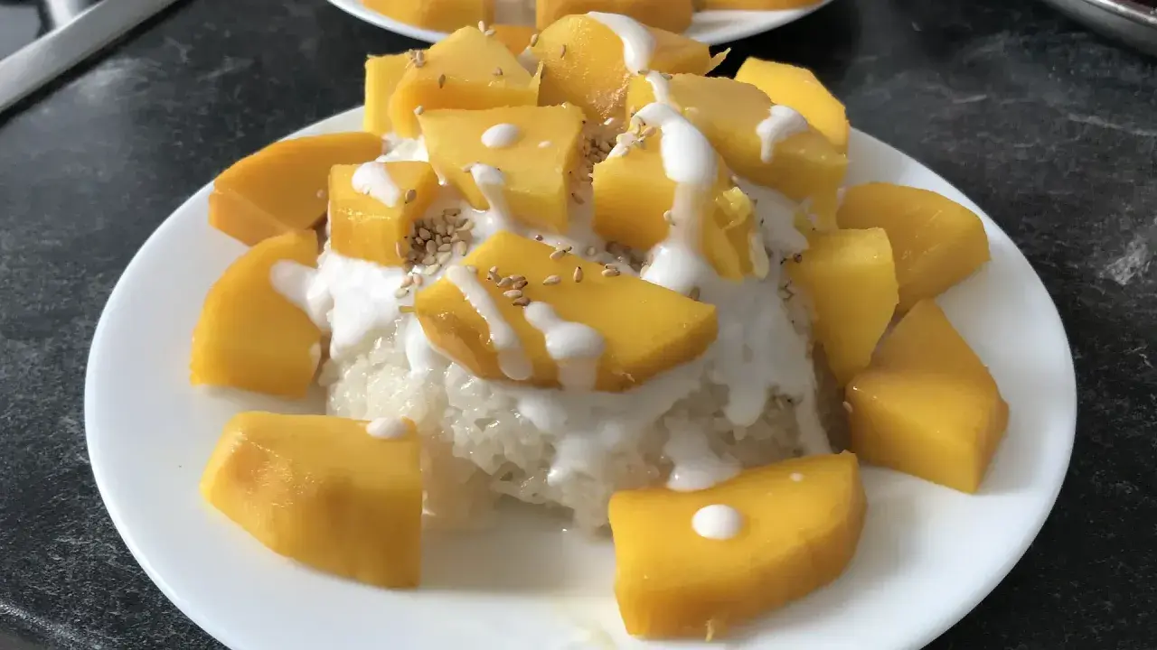 Ein Teller mit Klebreis, garniert mit saftigen Mango-Stücken, Kokosmilch und Sesam. Ein köstlicher **thai dessert mango**.