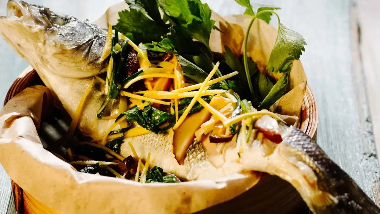 Gedämpfter Fisch, asiatisch zubereitet, mit Ingwer, Frühlingszwiebeln und frischen Kräutern in einem Bambuskorb.