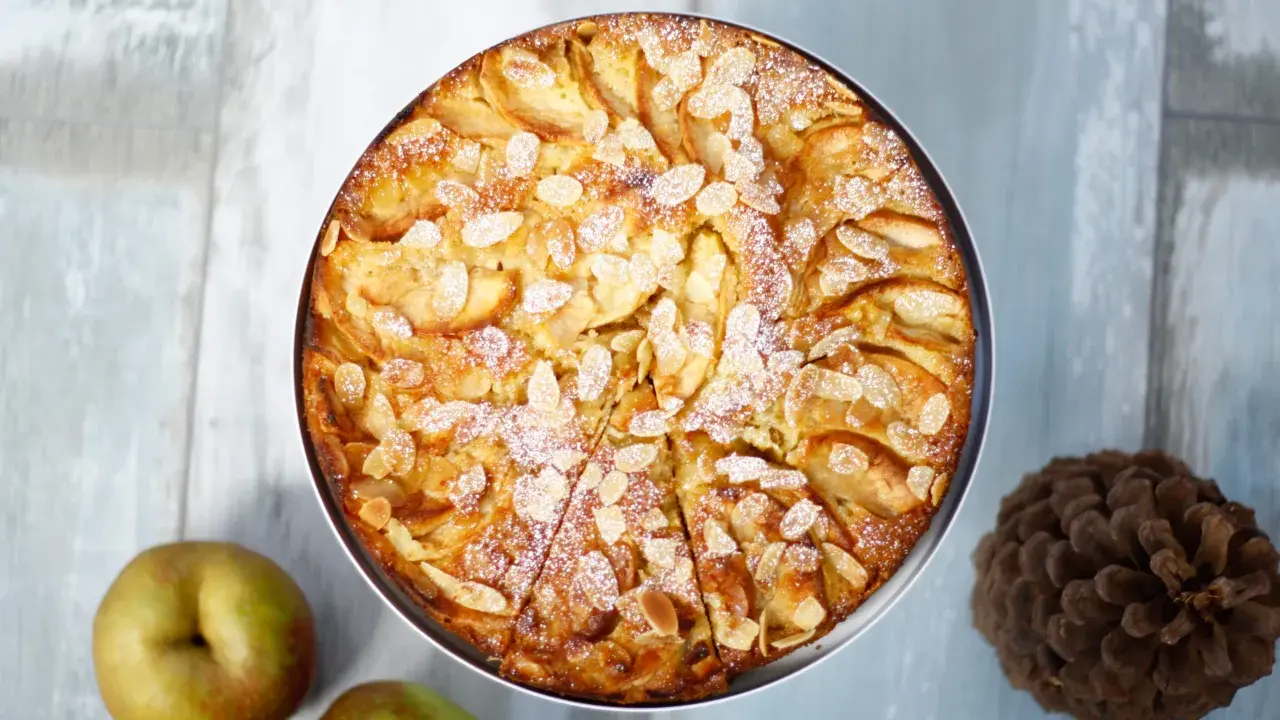 Ein goldbrauner bretonischer Apfelkuchen, bestreut mit Mandelblättchen und Puderzucker, neben Äpfeln und einer Kiefernzapfen.