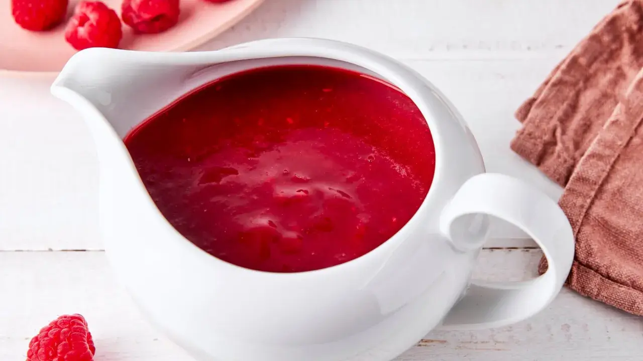 Himbeer coulis in einer weißen Soßenkelle, daneben frische Himbeeren und eine rosa Platte mit weiteren Früchten.