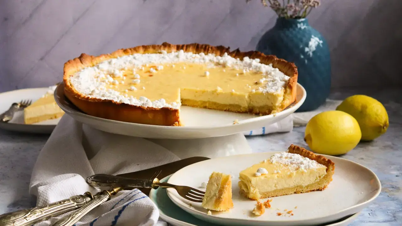 Eine köstliche Zitronen-Tarte, bestäubt mit Puderzucker, steht auf einem Kuchenständer. Ein Stück liegt auf einem Teller, daneben zwei frische Zitronen.
