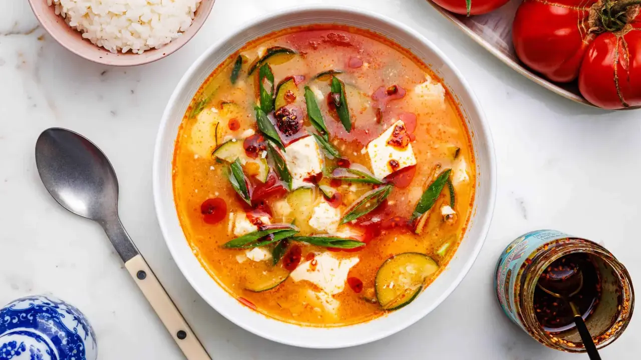 Eine dampfende Schüssel seidentofu suppe mit Zucchini und Frühlingszwiebeln, garniert mit Chiliöl. Daneben eine Schüssel Reis und ein Glas Chiliöl.