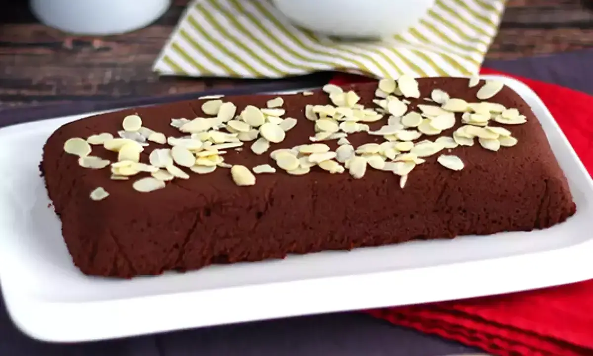 Dunkle, rechteckige marquise au chocolat, bestreut mit Mandelblättchen, auf einem weißen Teller.