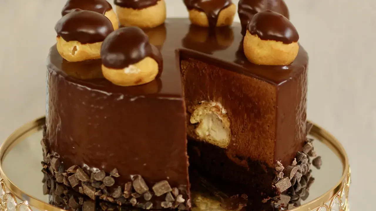 Eine glänzende Schokoladentorte, dekoriert mit kleinen Schokoladen-Profiteroles. Ein Stück ist herausgeschnitten und zeigt die cremige Füllung.