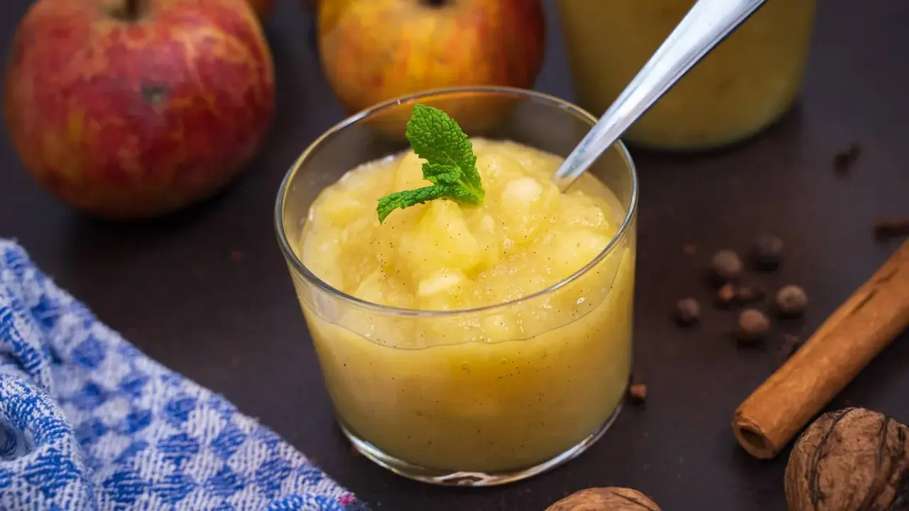 Ein Glas mit hausgemachtem compote de pommes, garniert mit Minze. Äpfel, Zimt und Nüsse im Hintergrund.