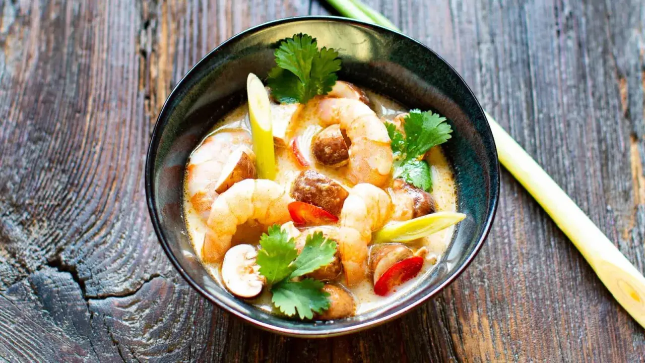 Eine Schüssel mit cremigem Tom Kha Gung, garniert mit Garnelen, Pilzen und Koriander, auf rustikalem Holz.