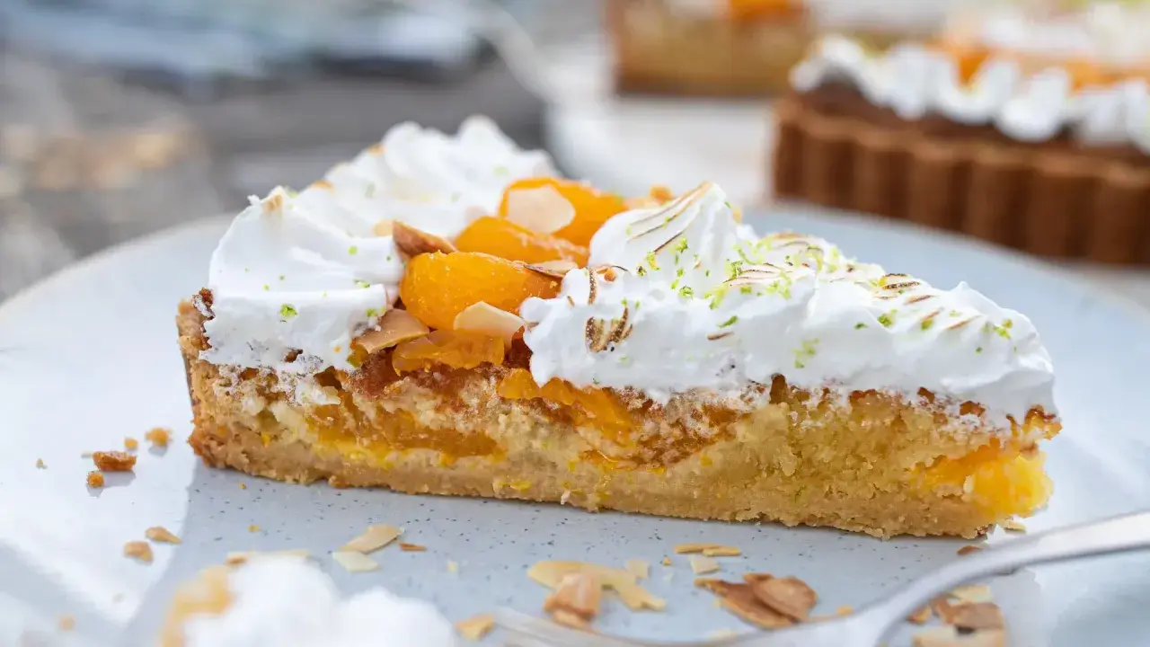 Ein Stück Mandarinen Tarte mit Baiserhaube, Mandelsplittern und frischer Zitronenschale bestreut.