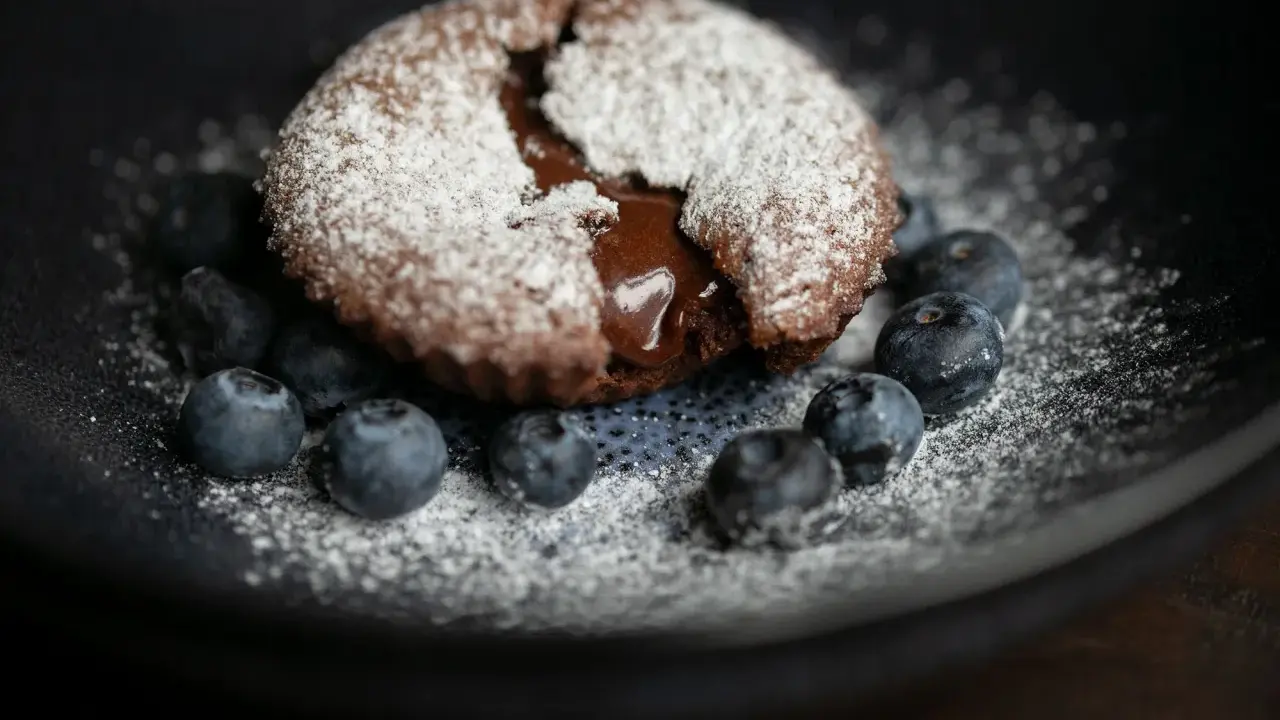 Ein saftiger Moelleux au Chocolat, bestäubt mit Puderzucker, umgeben von frischen Blaubeeren auf einem dunklen Teller.