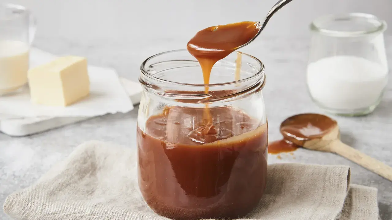 Ein Glas mit glänzender karamellsauce mit milch statt sahne. Ein Löffel hebt die cremige Sauce an.