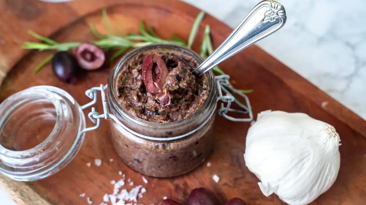 Ein Glas hausgemachte Tapenade, perfekt für Ihr nächstes Tapenade Rezept. Oliven, Knoblauch und Rosmarin auf einem Holzbrett.