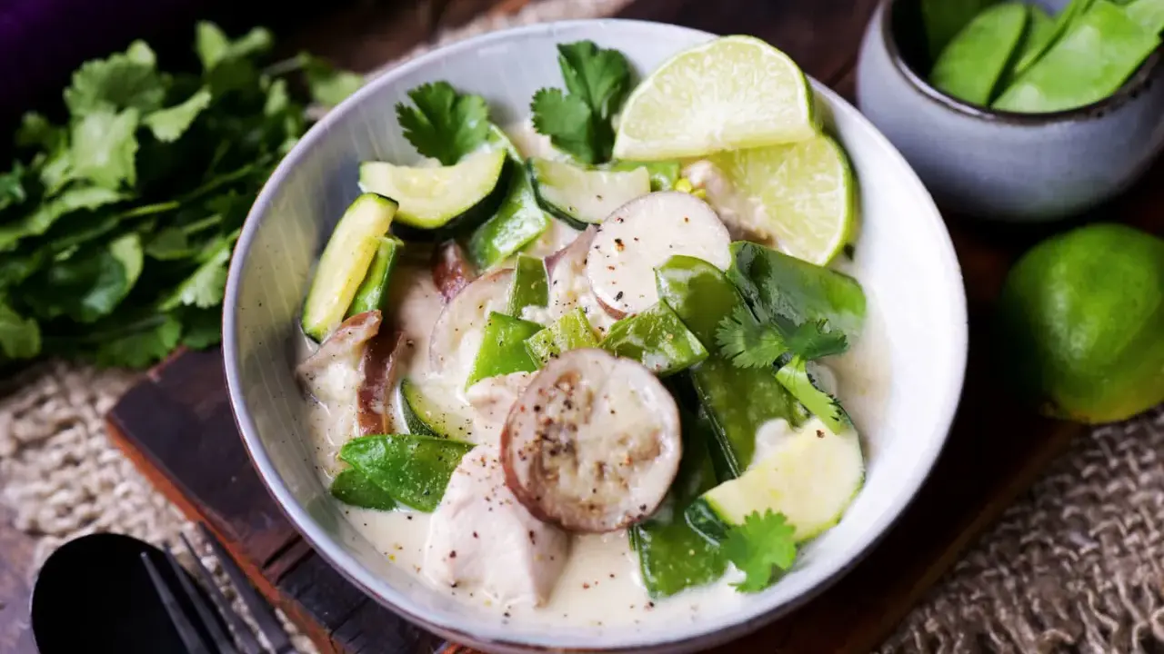 Ein cremiger Thai Green Curry mit Hühnchen, Zucchini, Zuckerschoten und Auberginenscheiben, garniert mit Limettenspalten und Koriander.