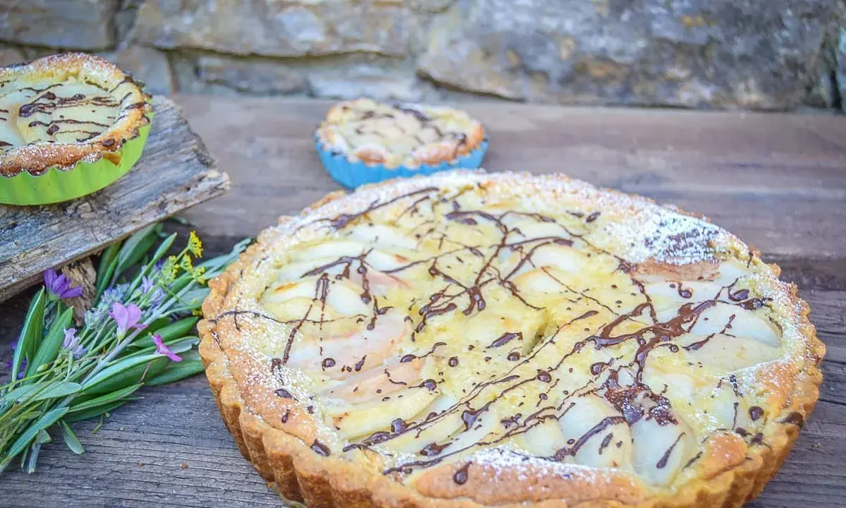 Ein köstlicher Amandine-Kuchen mit Birnenspalten und Schokoladendrizzle, bestäubt mit Puderzucker. Daneben kleine Törtchen und Kräuter.