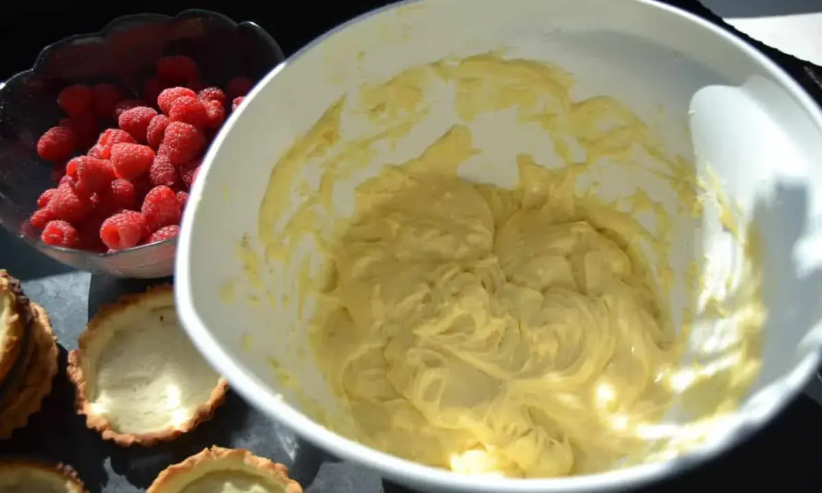 Frische Himbeeren warten neben leeren Tartelettförmchen und einer Schüssel mit cremiger Füllung auf die Fertigstellung der Himbeer Tartelettes.
