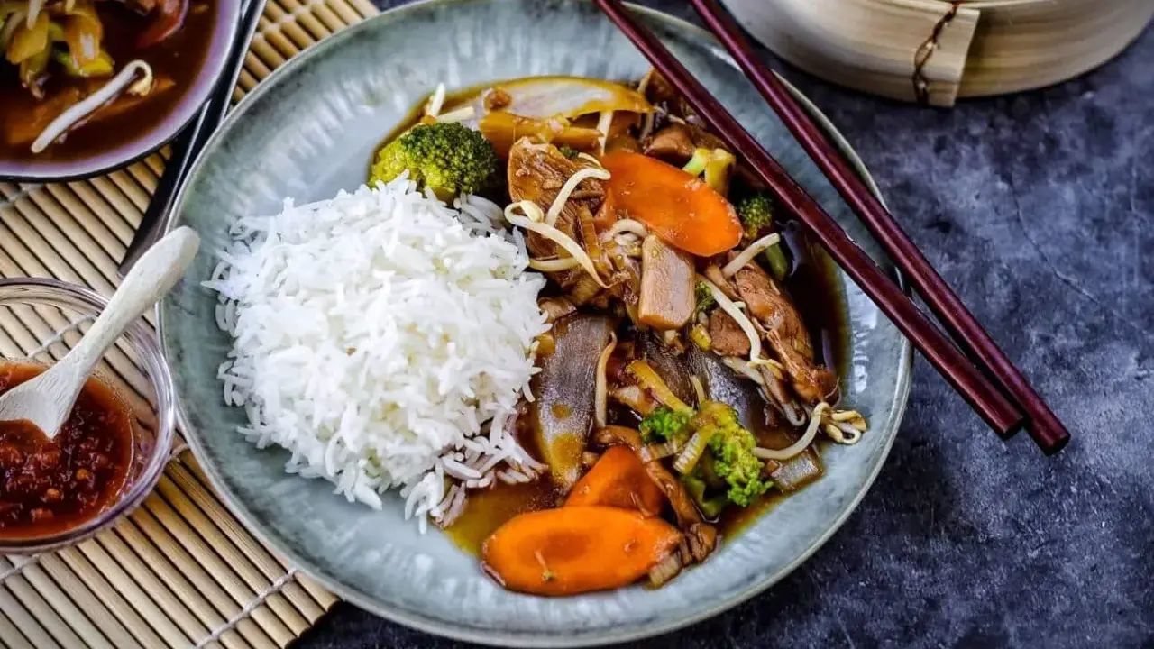 Reis mit einem herzhaften Stir-Fry-Gericht, das mit viel Gemüse und Fleisch in einer köstlichen Sauce serviert wird. Dazu gibt es eine kleine Schale mit scharfer Sauce.