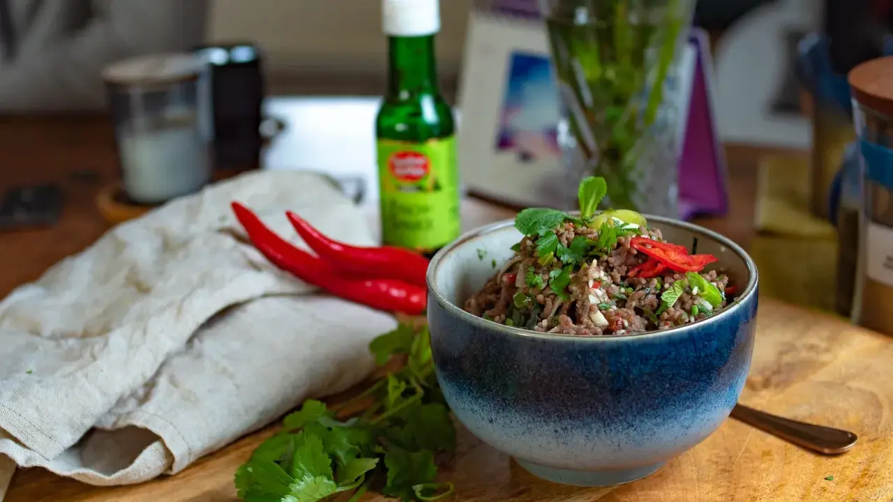 Schüssel mit würzigem Larb Moo, garniert mit frischen Kräutern und Chilis. Daneben liegen rote Chilis und eine Flasche Sauce.