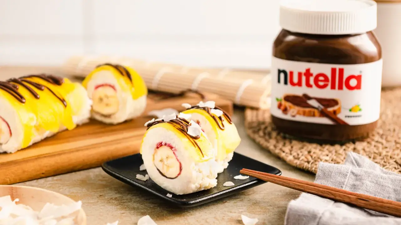 Süße Sushi-Rollen mit Milchreis als Basis, gefüllt mit Banane und Erdbeere, garniert mit gelbem Fruchtpüree, Schokodrizzle und Kokoschips. Daneben eine Nutella-Packung.