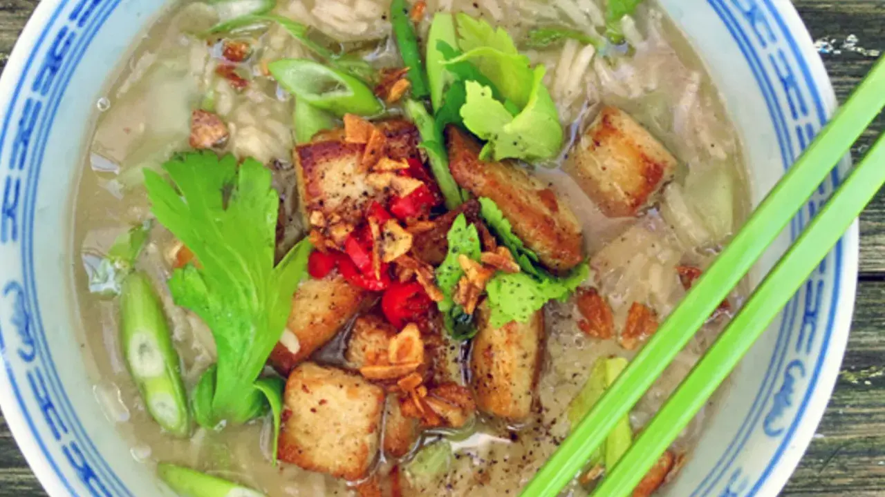 Khao tom, ein herzhafter Reissuppe mit gebratenem Tofu, Frühlingszwiebeln und Kräutern, serviert in einer blauen Schüssel mit Essstäbchen.