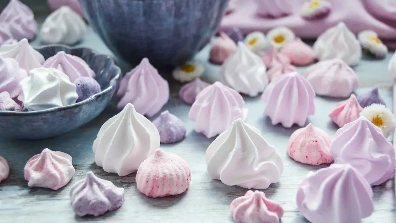 Zuckergebäck, was ist Meringue? Zarte, luftige Baisers in Rosa-, Lila- und Weißtönen, einige in einer Schüssel, andere verstreut.