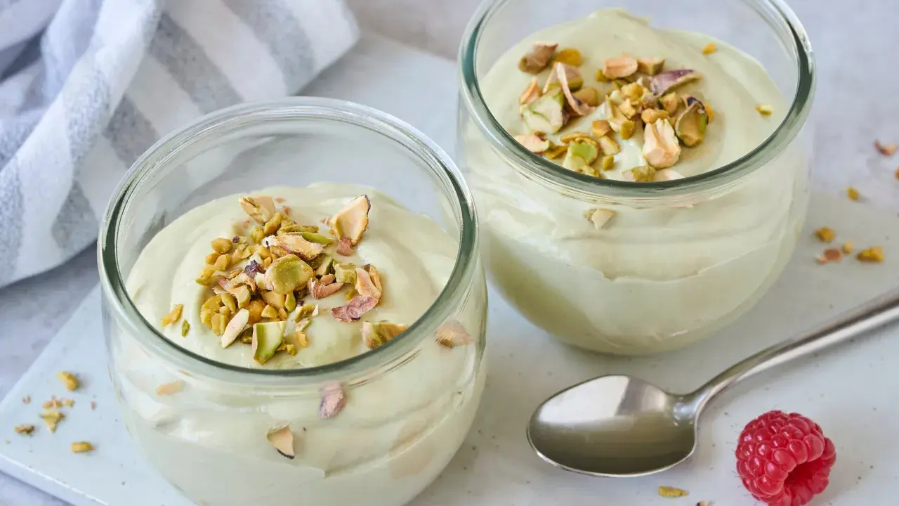 Zwei Gläser mit cremiger Pistazienmousse, garniert mit gehackten Pistazien. Ein Löffel und Himbeeren liegen daneben.