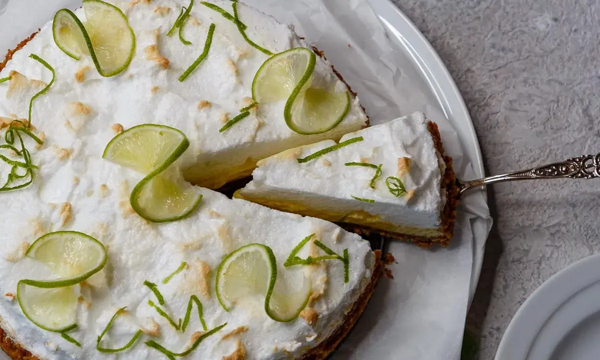 Eine köstliche Limetten Tarte mit luftiger Baiserhaube, garniert mit frischen Limettenscheiben und Zesten. Ein Stück wird gerade serviert.