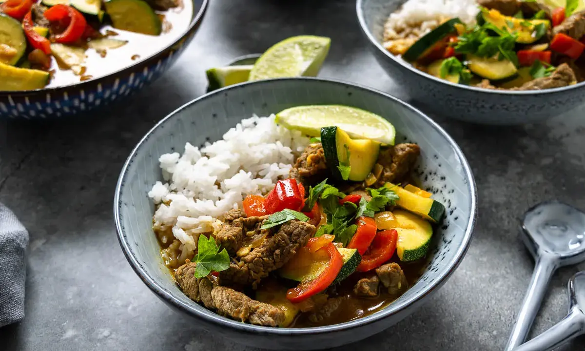Schüssel mit Reis und rotem Thai Curry mit Rindfleisch, Zucchini und Paprika, garniert mit Limettenspalten und Koriander.