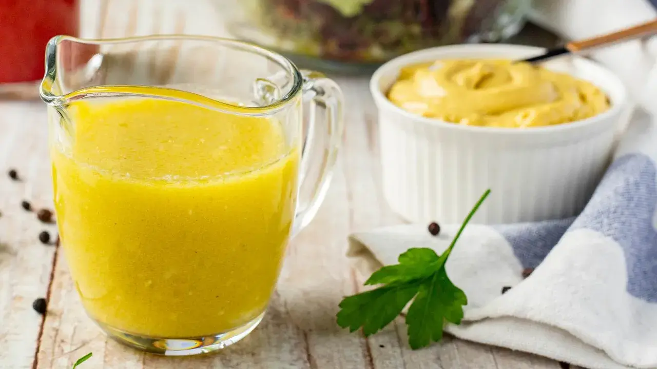 Gelbe Vinaigrette in einem Glasbehälter, bereit für einen frischen Salat. Daneben eine Schüssel Senf und ein Salat im Hintergrund.