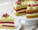 Eine wunderschöne Pistazien Torte mit Himbeerschichten und frischen Beeren. Ein Stück ist auf einem Teller angerichtet.