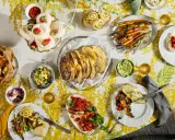 Tisch voller Oster-Essen-Ideen: Meringues mit Erdbeeren, Brot, Karotten, Quiche und mehr auf gelb-weißem Tischtuch.