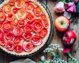 Ein wunderschöner Apfelrosen Kuchen, verziert mit zarten Apfelscheiben, die wie Rosen aussehen. Daneben liegen frische Äpfel und Blumen.