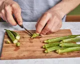 Hände schneiden Okra für ein leckeres Okra-Gericht. Frische grüne Schoten liegen auf einem Holzbrett.