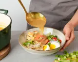 Eine goldene Schöpfkelle gießt eine cremige Brühe über Garnelen, Tofu, Ei und Sprossen in einer Schüssel. Ein köstliches Laksa Rezept wird zubereitet.