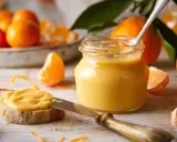 Ein Glas mit leuchtend gelbem orange curd steht neben frischen Orangen und einer Scheibe Brot, die damit bestrichen ist.