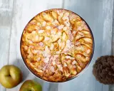 Ein goldbrauner bretonischer Apfelkuchen, bestreut mit Mandelblättchen und Puderzucker, neben Äpfeln und einer Kiefernzapfen.