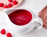 Himbeer coulis in einer weißen Soßenkelle, daneben frische Himbeeren und eine rosa Platte mit weiteren Früchten.