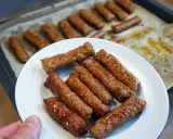 Leckere, glasiert aussehende Nem Nuong Würstchen auf einem Teller, weitere liegen auf einem Backblech.