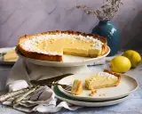 Eine köstliche Zitronen-Tarte, bestäubt mit Puderzucker, steht auf einem Kuchenständer. Ein Stück liegt auf einem Teller, daneben zwei frische Zitronen.