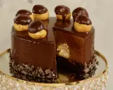 Eine glänzende Schokoladentorte, dekoriert mit kleinen Schokoladen-Profiteroles. Ein Stück ist herausgeschnitten und zeigt die cremige Füllung.