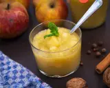 Ein Glas mit hausgemachtem compote de pommes, garniert mit Minze. Äpfel, Zimt und Nüsse im Hintergrund.