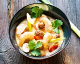Eine Schüssel mit cremigem Tom Kha Gung, garniert mit Garnelen, Pilzen und Koriander, auf rustikalem Holz.