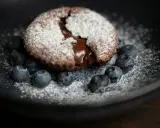 Ein saftiger Moelleux au Chocolat, bestäubt mit Puderzucker, umgeben von frischen Blaubeeren auf einem dunklen Teller.