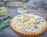 Ein köstlicher Amandine-Kuchen mit Birnenspalten und Schokoladendrizzle, bestäubt mit Puderzucker. Daneben kleine Törtchen und Kräuter.