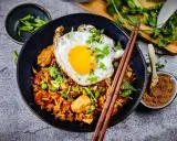 Was ist Nasi Goreng? Ein Teller mit gebratenem Reis, Hühnchen, Erbsen und einem Spiegelei, garniert mit Koriander und Frühlingszwiebeln.