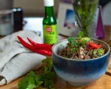 Schüssel mit würzigem Larb Moo, garniert mit frischen Kräutern und Chilis. Daneben liegen rote Chilis und eine Flasche Sauce.